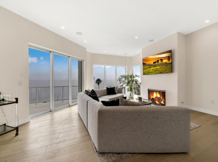 Angled view of living room space and fireplace. A large back of floor-to-ceiling windows and doors looking out to ocean