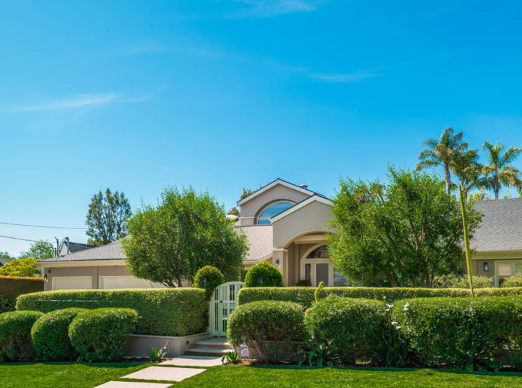 Large tan home with trimmed privacy hedges and palm trees