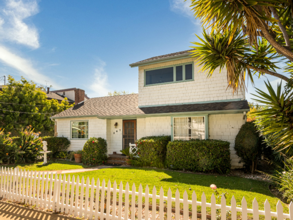 Two-story, cottage style, white-shingled house and front yard with hedges and palm trees