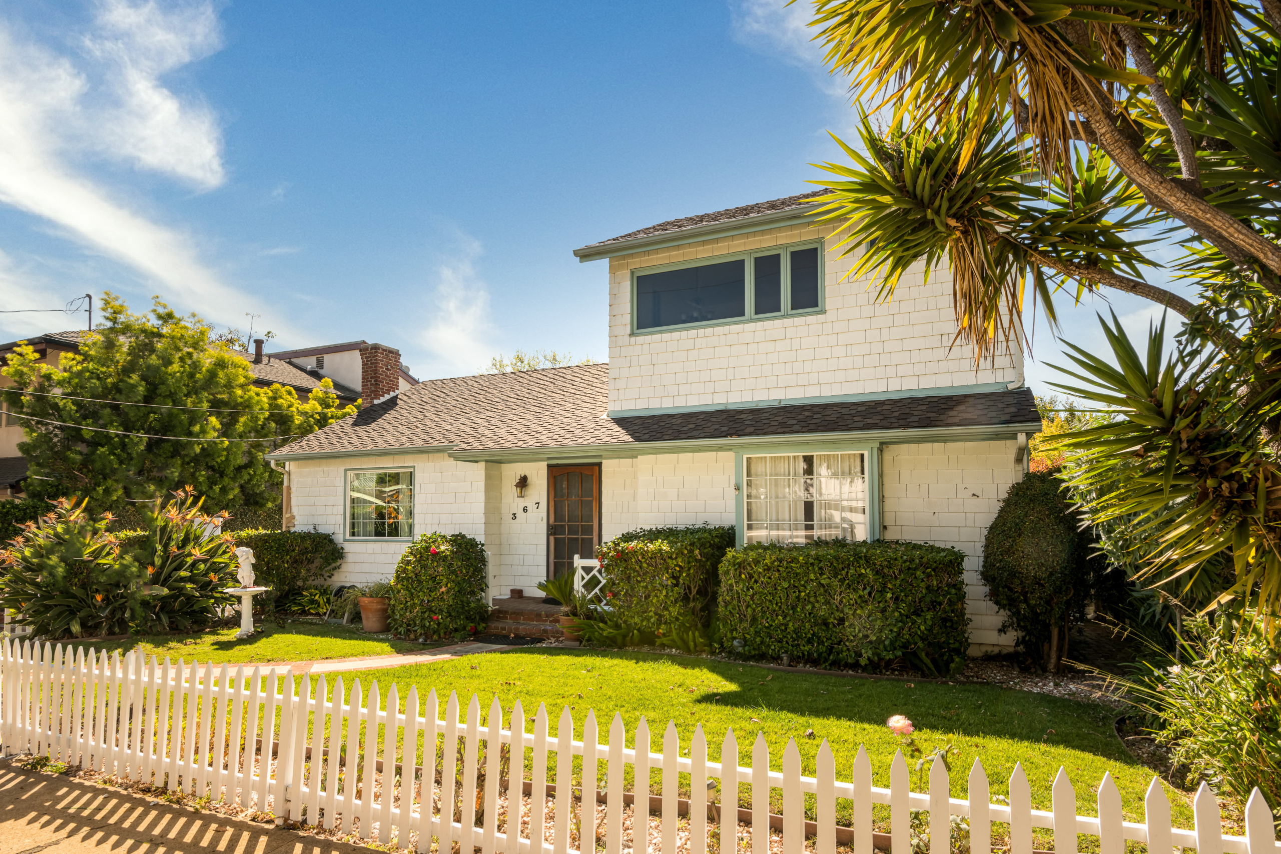 Two-story, cottage style, white-shingled house and front yard with hedges and palm trees