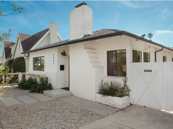 Front of California bungalow style home with white siding and brick chimney and gated driveway.