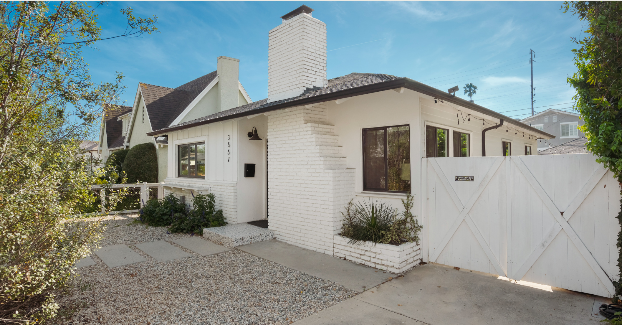 Front of California bungalow style home with white siding and brick chimney and gated driveway.